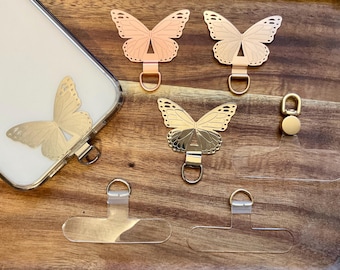 May include: A collection of butterfly-shaped phone charms in gold, rose gold, and silver tones, along with clear phone grip tabs. One charm is attached to a phone case. The charms have a loop for attaching to a phone.