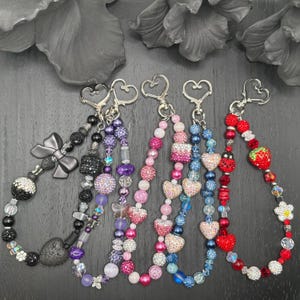 May include: A collection of beaded keychains in various colors. Each keychain features a heart-shaped clasp and is adorned with beads, crystals, and decorative charms, including hearts, bows, and flowers. The color palette includes pink, purple, red, and blue.