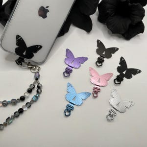 May include: A clear phone case with a black butterfly charm and a beaded lanyard. Several butterfly charms in various colors, including purple, pink, blue, and silver, are displayed. The charms have a metal clasp.