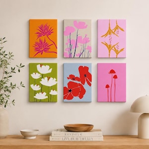 May include: Six framed floral art prints in vibrant colors. The artwork features stylized flowers in shades of pink, orange, red, and white against backgrounds of green, blue, and pink. The prints are displayed on a neutral wall.