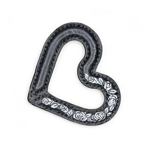 May include: A black heart-shaped decorative item with a textured surface and white floral accents. The item has a stitched edge and is set against a white background. The floral design includes roses and leaves.