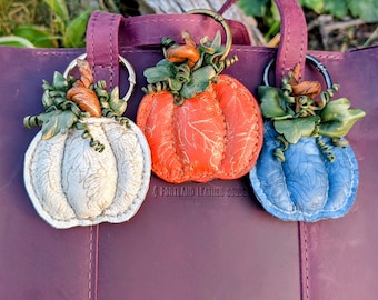 May include: Three leather pumpkin keychains in white, orange, and blue, each featuring a metallic ring, green leather leaves, and a brown stem. The pumpkins have a textured surface with a subtle leaf pattern and are attached to a purple leather bag.