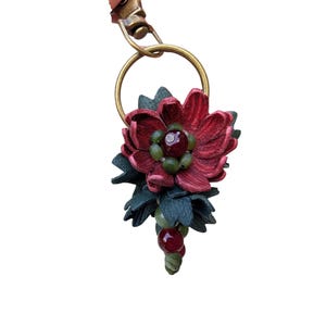 Op de afbeelding: Een handgemaakte bloemenhanger met een dieprode bloem, groene bladeren en een cluster van groene en rode kralen. De hanger is bevestigd aan een goudkleurige ring en een roze leren band. Het ontwerp is gedetailleerd en artistiek.