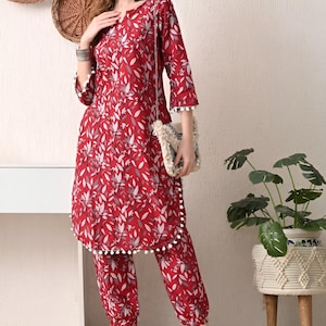 May include: A red floral print two-piece outfit. The top has three-quarter sleeves and white pom-pom trim. The matching pants have a similar floral pattern and pom-pom trim. A small, cream-colored purse with a strap is visible.