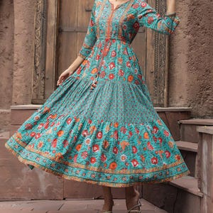 Peut inclure: Une longue robe turquoise à motifs floraux orange et rouges. La robe présente un col en V, des manches trois-quarts et une jupe à volants. Un cordon de serrage cintre la taille. La robe est portée avec des sandales dorées.