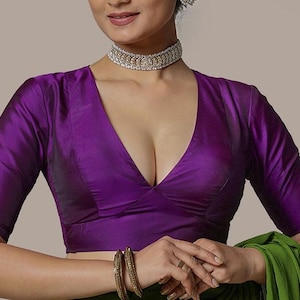 Könnte beinhalten: Eine tiefviolette Bluse mit tiefem Ausschnitt und ellenbogenlangen Ärmeln. Die Bluse hat einen seidigen Glanz und wird mit einem silbernen Choker und goldenen Armreifen kombiniert. Ein grüner Stoff ist darunter drapiert.