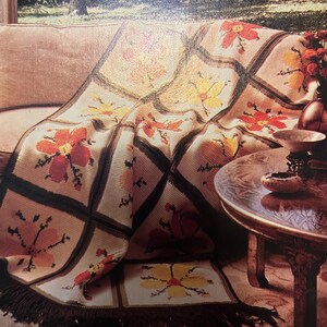May include: A beige crocheted blanket with a floral pattern in shades of red, orange, and yellow. The blanket features a grid design with dark brown borders and a fringed edge. The blanket is draped over a sofa.