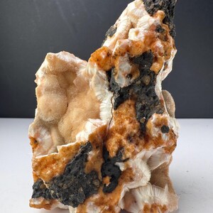 Puede incluir: Un espécimen mineral que presenta un grupo de cristales fibrosos blancos radiantes, intercalados con parches de roca volcánica texturizada de color gris oscuro y áreas de depósitos minerales cristalinos de tonos anaranjados.