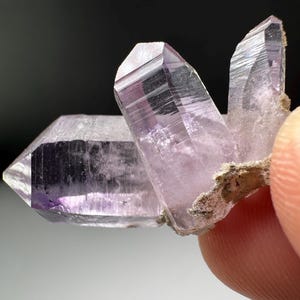 May include: A cluster of three translucent, light purple amethyst crystals with sharp, pointed terminations. The crystals exhibit natural facets and internal clarity, with a small amount of earthy, brownish matrix attached to the base of the cluster.