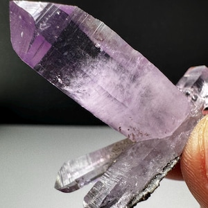 May include: A cluster of translucent, light purple amethyst crystals with sharp, pointed terminations. The crystal formation features distinct vertical striations along the facets and a dark, textured mineral base where the points converge.