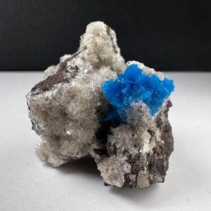 May include: A mineral specimen featuring a cluster of vibrant, electric blue crystals embedded within a rough, textured matrix of translucent white and clear crystalline formations, set against a dark, solid background.