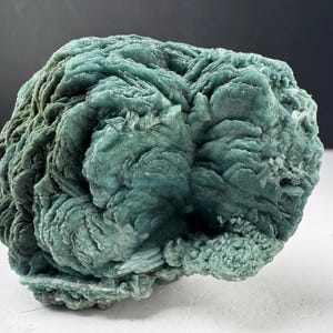 May include: A raw mineral specimen featuring a dense, rounded cluster of layered, plate-like formations in shades of teal and sage green. The surface displays a complex, swirling texture with visible crystalline edges and a matte finish throughout.