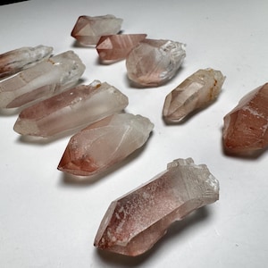 May include: A collection of raw, natural quartz crystals. The crystals vary in size and shape, with a clear to translucent appearance and reddish-brown inclusions. They are arranged on a white surface, showcasing their unique textures and colors.