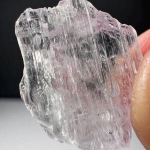 May include: A translucent, pale pink kunzite crystal specimen with a distinct striated, vertical texture across its surface. The mineral fragment features jagged, irregular edges and a clear, glass-like appearance with internal light reflections.