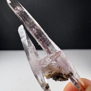 May include: A translucent, light purple amethyst crystal cluster featuring two elongated, pointed terminations extending from a central base with dark, earthy mineral inclusions visible at the connection point of the crystal structure.
