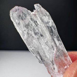 May include: A translucent, pale pinkish-white crystal specimen with a jagged, elongated shape and a striated, fibrous surface texture. The mineral displays natural fractures and internal clarity, with a light-catching, glassy luster throughout its structure.