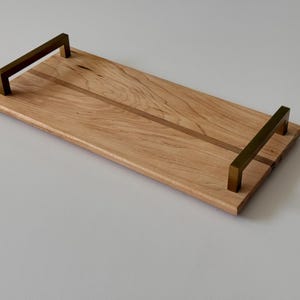 May include: A rectangular wooden serving tray with a light wood grain pattern. It features two rectangular brass handles, one on each end. The tray is approximately 18 inches long and 9 inches wide.