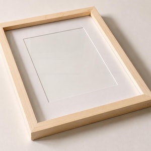 May include: A rectangular picture frame with a light-colored natural wood finish. The frame features a clean, simple design with mitered corners and includes a white mat board centered within the frame, providing a clear display area for artwork.