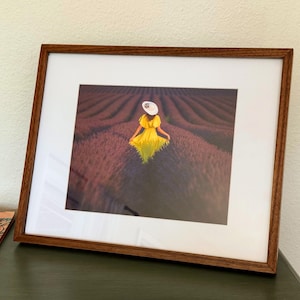 May include: A framed photograph featuring a person in a yellow dress and white hat walking through a lavender field. The photograph is set within a brown wooden frame with a white mat, resting on a dark green surface.