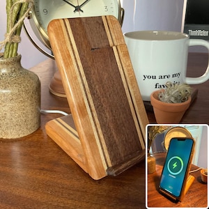 May include: A wooden wireless phone charger with a striped design in shades of brown and tan. The charger is angled, with a phone displaying a charging symbol. A white mug with the text "you are my favorite" is in the background.