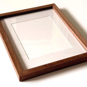 May include: A rectangular picture frame constructed from medium-toned brown wood with a visible grain pattern. The frame features a clean, flat profile and includes a crisp white mat board centered within the frame, surrounding a blank white space.