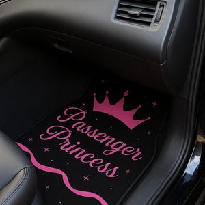 May include: Black car floor mat with the words "Passenger Princess" in pink script, a pink crown, and star accents. The mat is placed in the passenger side of a car.
