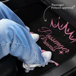 May include: A black car floor mat featuring a pink graphic of a crown and the text "Passenger Princess" in a cursive font. The mat includes a small star icon and the additional text "Passenger Princess approved" with a white arrow pointing down.