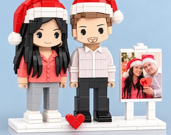 May include: Two custom brick-built figures wearing red and white Santa hats, standing on a white base with a small red heart. One figure has long black hair and a pink shirt, the other has brown hair and a white shirt. A framed photo stands beside them.
