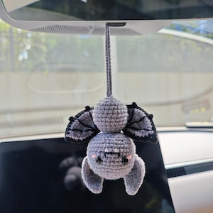 Può includere: Un ciondolo a forma di pipistrello all'uncinetto, appeso allo specchietto retrovisore di un'auto. Il pipistrello è grigio con dettagli neri sulle ali e un viso sorridente. Il ciondolo è attaccato da un cordoncino all'uncinetto grigio.
