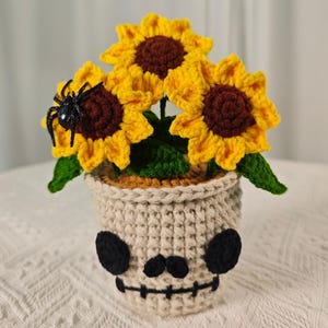May include: Crocheted sunflower arrangement in a skull-shaped pot. The sunflowers have yellow petals and brown centers, with green leaves. A black spider sits on one flower. The pot is beige with black eye and mouth details.