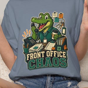 Può includere: Una t-shirt blu ardesia con la grafica di un alligatore verde seduto a una scrivania con un telefono, una tazza di caffè e documenti. Il testo "FRONT OFFICE CHAOS" è stampato in grassetto verde e bianco sotto l'illustrazione.