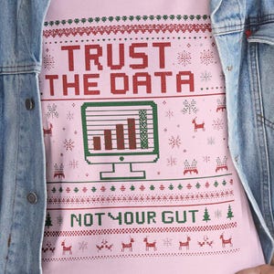 Può includere: T-shirt rosa girocollo con la scritta "TRUST THE DATA... NOT YOUR GUT" in rosso e verde. Il design include una grafica pixelata dello schermo del computer e motivi festivi di fiocchi di neve e renne.