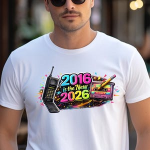 Può includere: T-shirt bianco con un design grafico colorato con un telefono cellulare retrò, una musicassetta e la scritta "2016 is the New 2026". Il design ha una vivace combinazione di colori neon.