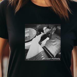 Può includere: T-shirt nera con un'immagine in bianco e nero di una persona in ginocchio. Il testo "Kneel Politely" è visibile in basso nell'immagine. La maglietta è indossata da una persona.