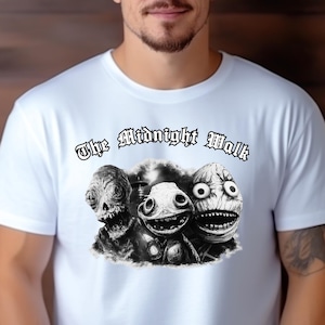 May include: White t-shirt featuring a black and white graphic of three stylized monster faces with large eyes and open mouths. The text "The Midnight Walk" is arched above the graphic in a gothic font.