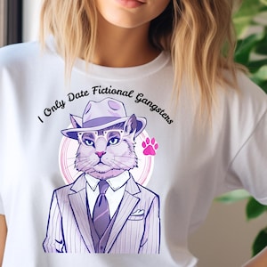 Può includere: T-shirt bianca con un gatto dei cartoni animati vestito con un abito da gangster e un fedora. Il gatto è all'interno di un cerchio rosa con un'impronta di zampa. Il testo sulla maglietta recita "I Only Date Fictional Gangsters."