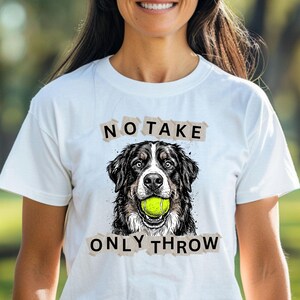 Può includere: T-shirt bianca con una grafica di un cane che tiene una pallina da tennis. Il testo "NO TAKE ONLY THROW" è stampato sopra e sotto l'illustrazione del cane. La maglietta viene indossata all'aperto.