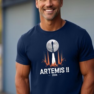 Può includere: T-shirt blu navy con grafica di un razzo che si lancia verso la luna piena, con la scritta "ARTEMIS II - 2026" in bianco. Il design include fiammate arancioni e bianche. La maglietta è a maniche corte.