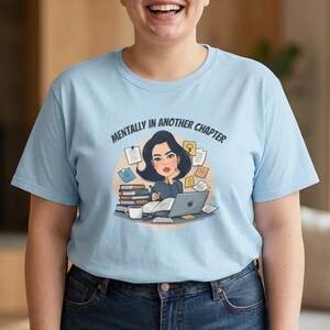 Può includere: Una maglietta azzurra con la scritta "Mentally in Another Chapter" e una grafica di una donna che lavora su un laptop, circondata da libri e fogli. La persona nell'immagine indossa la maglietta e jeans blu.