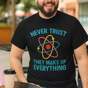 Può includere: T-shirt nero con la scritta "NEVER TRUST THEY MAKE UP EVERYTHING" e una grafica colorata di un atomo. La maglietta è indossata da una persona seduta a un tavolo con altre due persone. La maglietta è in materiale morbido.