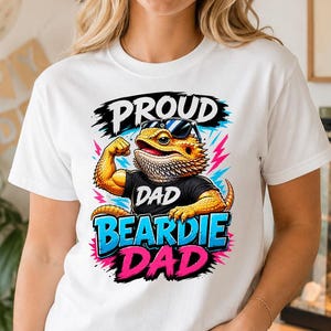 Può includere: T-shirt bianca con la scritta "PROUD DAD BEARDIE DAD" e un'illustrazione a cartoni animati di un drago barbuto con occhiali da sole e che flette il braccio. La maglietta è indossata da una persona.