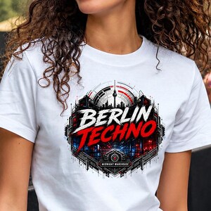 May include: White t-shirt with a graphic that says "Berlin Techno" in red and white. The design includes an image of the Berlin skyline and a geometric border. The shirt is being worn outdoors.