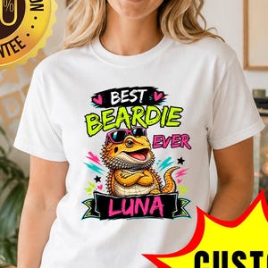 Può includere: T-shirt bianca con una grafica colorata di un drago barbuto dei cartoni animati con occhiali da sole e la scritta "BEST BEARDIE EVER LUNA". Sullo sfondo un sigillo dorato "SODDISFAZIONE DEL CLIENTE 100% GARANTITA".