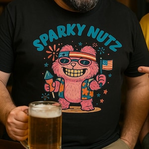 May include: Black t-shirt with the text "SPARKY NUTZ" in blue and a cartoon bear wearing sunglasses, holding a firework and an American flag. The bear is pink and wearing a floral jacket. A person is holding a beer mug.