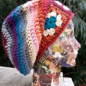 以下が含まれることがあります： 花柄のカラフルな手編みのビーニー帽。帽子は、紫、青、ターコイズ、グレー、ピンク、オレンジ、赤など、さまざまな色で作られています。帽子はマネキンの頭に載っています。