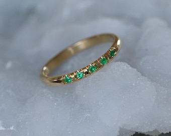 May include: A delicate gold band ring with a row of small, green emerald gemstones.
