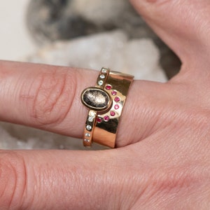 K&ouml;nnte beinhalten: Ein Set aus drei goldenen Ringen. Ein Ring hat einen ovalen, dunkel gef&auml;rbten Stein. Ein weiterer Ring hat kleine, runde, rote Steine. Der dritte Ring hat kleine, klare Steine. Die Ringe sind aufeinander gestapelt.