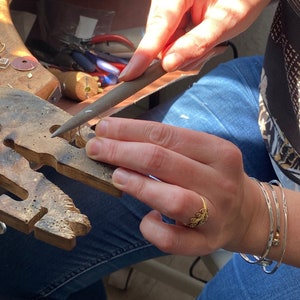K&ouml;nnte beinhalten: Eine Person arbeitet an Schmuck und verwendet eine Feile. Die Person tr&auml;gt einen goldenen Ring und silberne Armb&auml;nder. Der Arbeitsbereich umfasst Werkzeuge und eine Holzwerkbank. Die Person tr&auml;gt eine blaue Jeans.
