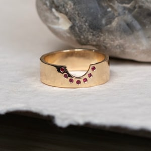 May include: A gold ring with a flat band and a curved cutout. The ring features seven small, round, red gemstones set along the curved edge. The ring is displayed on a white surface.