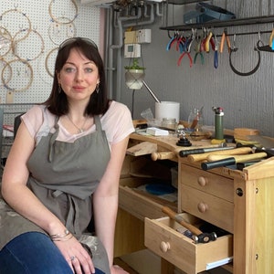 May include: A woman wearing a gray apron and a white shirt is standing in front of a workbench with tools and jewelry making supplies. The workbench has drawers and a wooden top.